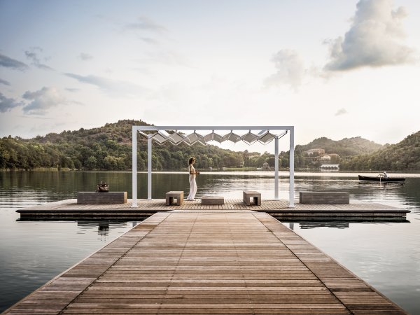 Die freistehende Pergola Gibus Med Open Fly mit beweglichen Dach auf einem idyllischen Holzsteg am Wasser.