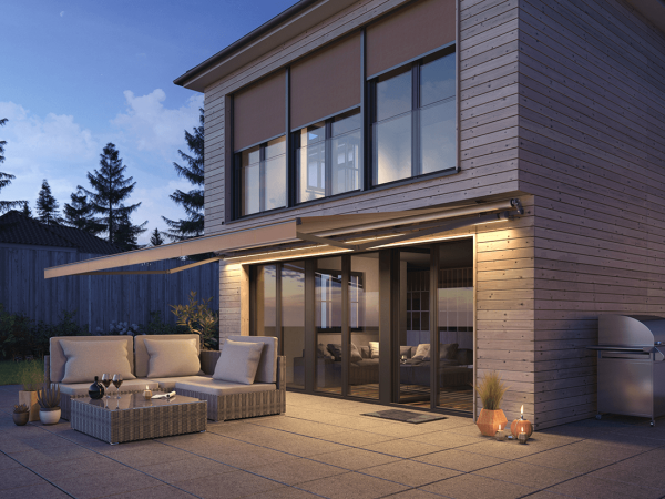 Die offene Gelenkarmmarkise von Frimey an einem modernen Holzhaus mit Lounge Outdoor Möbel auf der Terrasse.